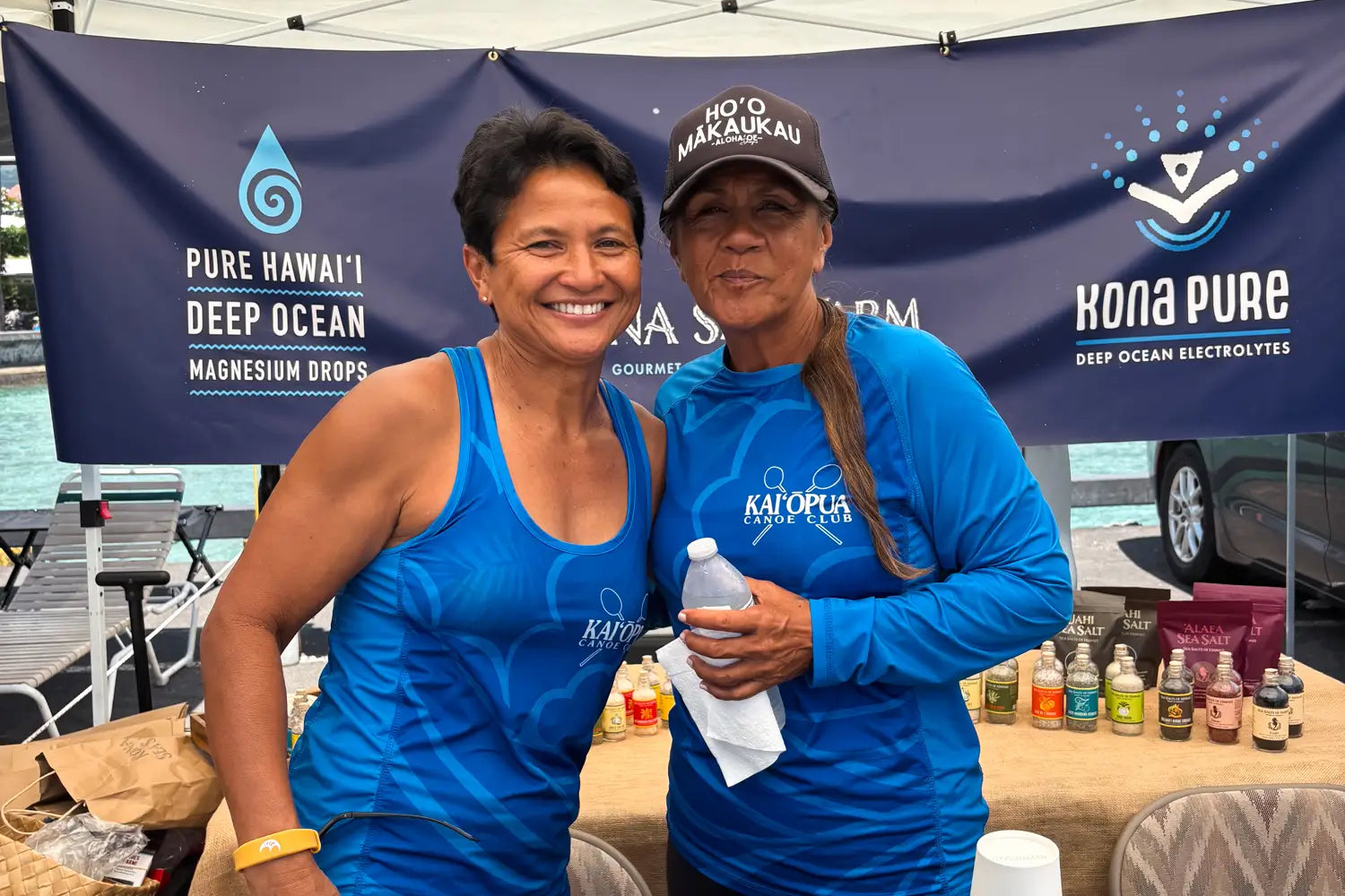 Two women in blue shirts standing in front of a Kona Pure banner at an outdoor event.
