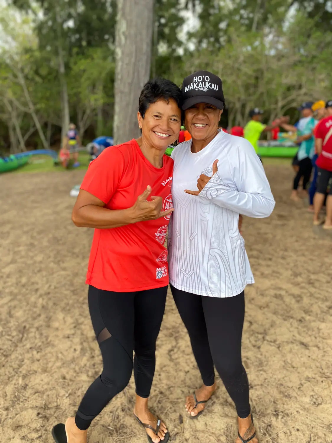 Two women posing together outdoors with trees and people in the background